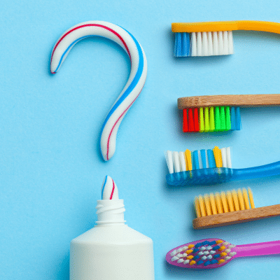 A group of toothbrushes next to a tube of toothpaste with paste squeezed out to make a question mark, representing how to choose a toothbrush and toothpaste